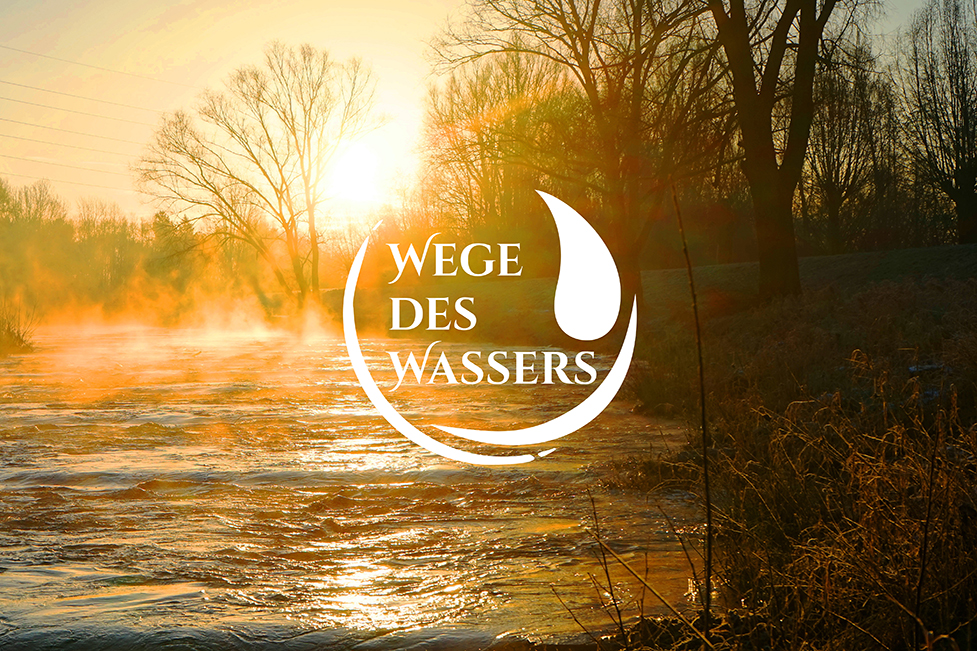 Ein wunderschöner Blick auf die Vechte bei Sonnenaufgang. Leichter Nebel steigt vom Fluß auf, der in goldenes Licht getaucht ist. In der Mitte des Bildes ist das Logo "Wege des Wassers" zu sehen.