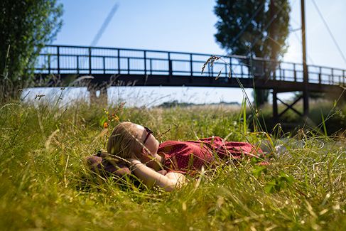 Eine junge Frau liegt bei Sonnenschein gemütlich im Gras. Im Hintergrund ist eine Holzbrücke zu sehen.