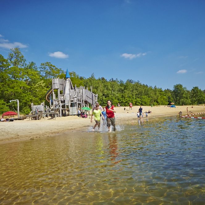 Blick auf den See und den Kletterturm, eine Familie tollt am Wasser herum und genießt sichtlich das schöne Wetter.