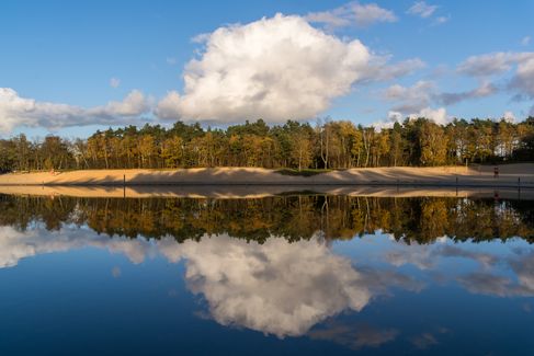 Ufer, blauer Himmel und Wolken spiegeln sich im stillen Wasser des Quendorfer Sees.