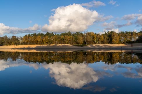 Vom anderen Ufer blicken wir auf den Sandstrand des Quendorfer Sees. Bäume und Wolken spiegeln sich im ruhigen Wasser.