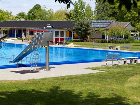 Durch Sträucher hindurch ist das Freibad in Schüttorf im Sonnenschein zu sehen.