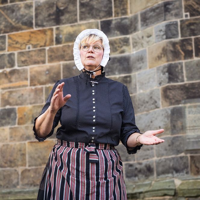 Ein Dame in traditioneller Kleidung, mit Haube und Schürze, erklärt etwas. Im Hintergrund sehen wir das Mauerwerk des Historischen Rathauses.