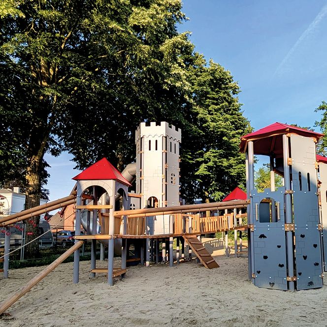 Die Sonne scheint auf den von grünen Bäumen umrandeten Ritterspielplatz, auf dem die Türme und eine kleine Hängebrücke im Vordergrund zu sehen sind.